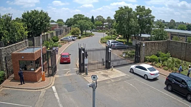 Gate camera: a guard at the gatehouse, the vehicle gate open with a red car passing through, two people on the pavement